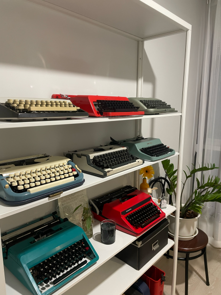Typewriters on three shelves in a variety of colors (red, teal, grey with white or black keys). 